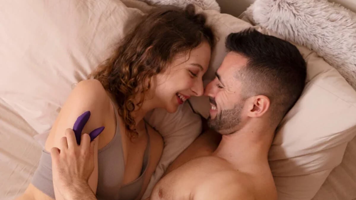 A young couple lies in bed smiling at each other, holding a sex toy in their hands
